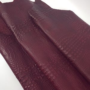May include: Close-up of burgundy-colored leather with a crocodile skin texture. The leather is folded and has a detailed, embossed pattern. The material appears to be high-quality and suitable for various crafting projects.