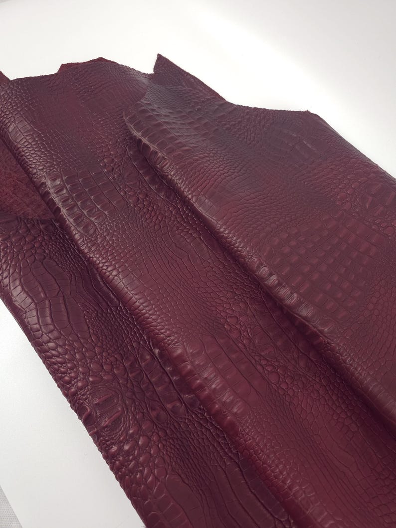 May include: Close-up of burgundy-colored leather with a crocodile skin texture. The leather is folded and has a detailed, embossed pattern. The material appears to be high-quality and suitable for various crafting projects.