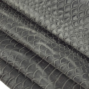 Könnte beinhalten: Grauer Kunstlederstoff mit einem strukturierten, schuppigen Muster. Der Stoff ist in mehreren Lagen gefaltet, wodurch die Textur aus verschiedenen Blickwinkeln sichtbar wird.