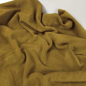 May include: Mustard-coloured fabric with a soft, suede-like texture. The fabric is draped and folded, creating shadows and highlights that emphasise its texture. The colour is a warm, earthy tone, and the fabric appears to be lightweight and flowing.
