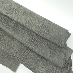 May include: A close-up of a folded piece of gray faux crocodile leather. The material has a textured surface with a repeating pattern that mimics the scales of a crocodile. The leather is a muted gray color.