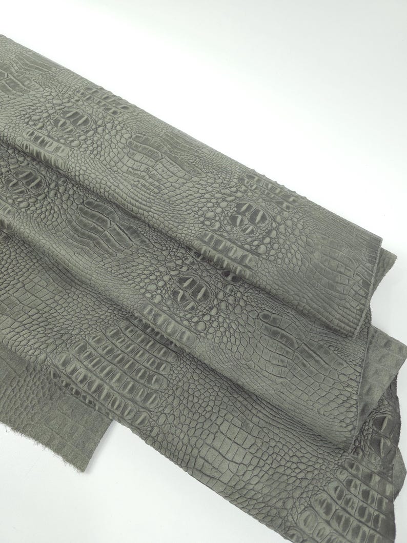 May include: A close-up of a folded piece of gray faux crocodile leather. The material has a textured surface with a repeating pattern that mimics the scales of a crocodile. The leather is a muted gray color.