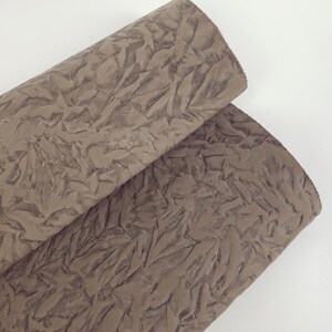 May include: Two rolls of brown fabric with a textured, wrinkled surface. The fabric has a matte finish and appears to be made of a soft material. The rolls are positioned diagonally against a white background.