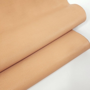 May include: A close-up of a folded piece of tan-colored leather. The leather has a textured, reptile-like pattern. The words "Leatherforcraft" are printed in white at the bottom left corner.