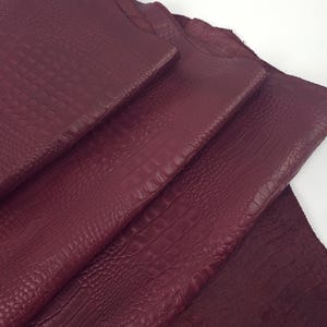 Op de afbeelding: Close-up van verschillende vellen bordeauxrood leer. Het leer heeft een textuur met een krokodilachtig patroon. De vellen zijn gelaagd en tonen de verschillende texturen en dieptes van het materiaal. De kleur is een diepe, rijke tint rood.