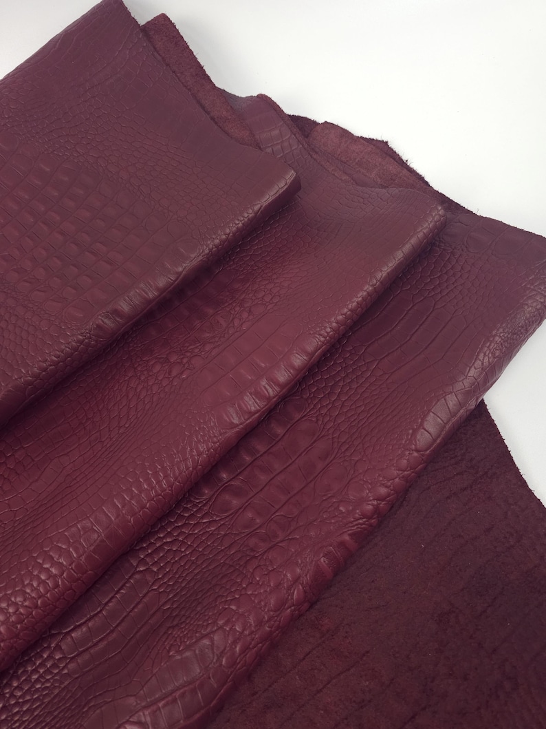 May include: Close-up of several sheets of burgundy-colored leather. The leather has a textured, crocodile-like pattern. The sheets are layered, showing the varying textures and depths of the material. The color is a deep, rich shade of red.