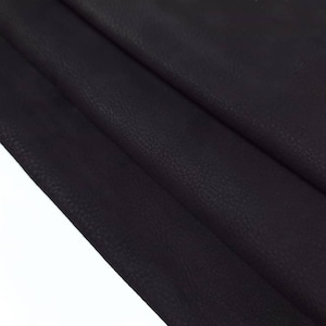 Può includere: Primo piano di un materiale in pelle marrone scuro con una superficie strutturata. La pelle è piegata, rivelando la grana e sottili variazioni di colore. Il materiale appare morbido e flessibile, adatto a vari progetti artigianali.