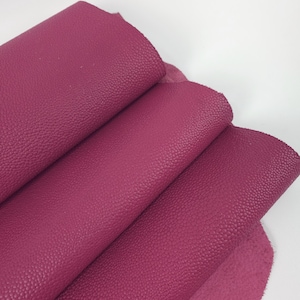 May include: Close-up of several sheets of deep magenta-colored leather. The leather has a textured, pebbled surface. The sheets are layered, revealing the underside of one sheet. The background is plain white.
