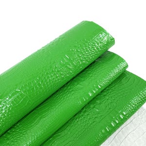May include: Bright green faux leather sheets with a crocodile skin texture. The sheets are rolled up and layered, showcasing the glossy finish and detailed pattern. The bottom right corner shows a glimpse of white leather. The text "leatherforcraft" is visible.
