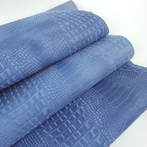 May include: A folded length of periwinkle blue fabric featuring a textured, embossed pattern that mimics the appearance of reptile skin. The material displays a repeating scale design with varying sizes of raised, rounded, and rectangular shapes.
