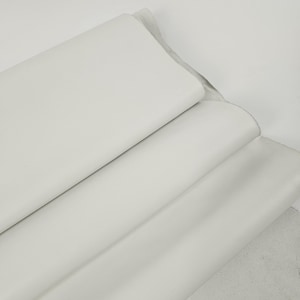 Puede incluir: Primer plano de varios rollos de cuero blanco. El cuero es liso y parece de alta calidad. Los rollos están apilados, con los bordes visibles. El fondo es blanco liso, resaltando el color y la textura del cuero.