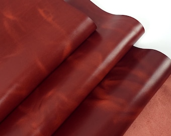 May include: Close-up of several sheets of burgundy-colored leather. The leather has a smooth, glossy finish with subtle variations in color and texture. The sheets are layered, revealing a lighter, reddish-brown underside.