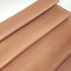 Puede incluir: Primer plano de una hoja de cuero sintético plegada con un patrón de piel de serpiente. El material es de color cobre cálido, con un brillo sutil. La textura de la piel de serpiente es visible, agregando profundidad y detalle.