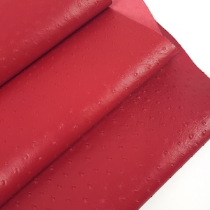 May include: Close-up of folded red leather with a textured surface. The leather has a consistent color and a pattern of small, evenly spaced bumps. The material appears to be soft and flexible, with a slight sheen.