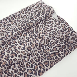 Könnte beinhalten: Ein gefaltetes Stück Stoff mit einem sich wiederholenden Leopardenmuster. Das Design besteht aus dunkelbraunen und schwarzen Flecken, die in unregelmäßigen Formen auf einem hellbeigen Hintergrund angeordnet sind und dem Textil ein strukturiertes Aussehen verleihen.