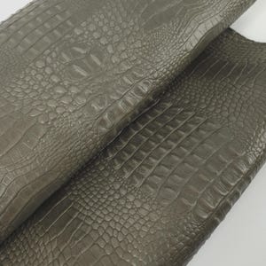 May include: Close-up of a roll of olive-green faux crocodile leather. The material has a textured surface with a scale pattern, and is folded over itself. The lighting highlights the texture and color variations.