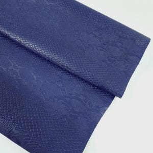 Peut inclure: Une feuille de similicuir bleu marine avec une texture peau de serpent. Le matériau est plié, révélant le dessous. La surface présente un motif en relief détaillé, créant une apparence réaliste de peau de reptile. Le fond est blanc uni.