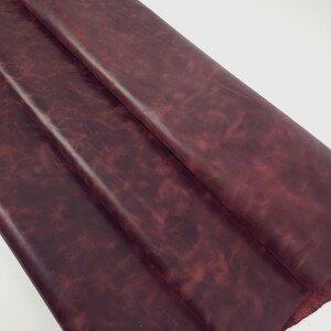 May include: A large, folded piece of deep burgundy leather with a smooth, matte finish. The surface displays a subtle, mottled texture with variations in color intensity, showing darker and lighter shades of red throughout the material.