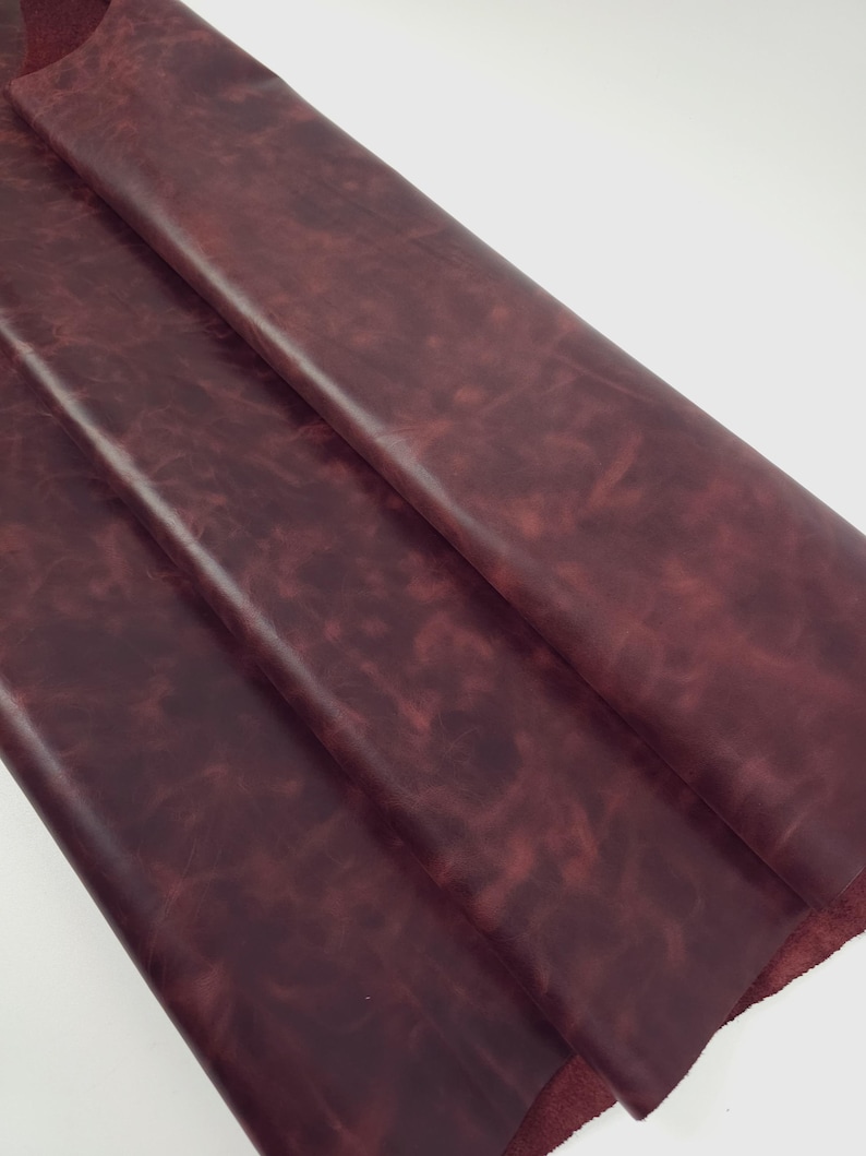May include: A large, folded piece of deep burgundy leather with a smooth, matte finish. The surface displays a subtle, mottled texture with variations in color intensity, showing darker and lighter shades of red throughout the material.