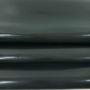 May include: Close-up of a dark green, glossy leather material. The leather is folded, revealing multiple layers and a smooth, reflective surface. The color is a deep, muted green.