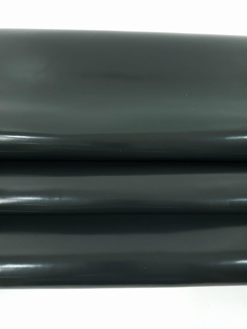 May include: Close-up of a dark green, glossy leather material. The leather is folded, revealing multiple layers and a smooth, reflective surface. The color is a deep, muted green.