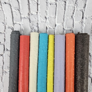 Könnte beinhalten: Neun Rollen aus Straußenleder mit Prägung in verschiedenen Farben, darunter Schwarz, Rot, Weiß, Blau, Gelb, Lila und Orange. Das Leder ist vertikal an einer weißen Backsteinwand angeordnet. Der Text "leatherforcraft" ist am unteren Rand des Bildes sichtbar.