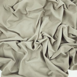 Pode incluir: Um close-up de um grande pedaço de tecido em um tom neutro e suave. O material é drapeado e dobrado, criando sombras e destaques. O tecido tem uma aparência ligeiramente texturizada, sugerindo uma sensação macia e luxuosa.