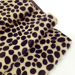 Può includere: Un pezzo di pelle animale caratterizzato da una base color beige naturale con un fitto motivo di macchie circolari dal marrone scuro al nero. La consistenza appare corta e pelosa, con il materiale ripiegato per mostrare il lato inferiore marrone.