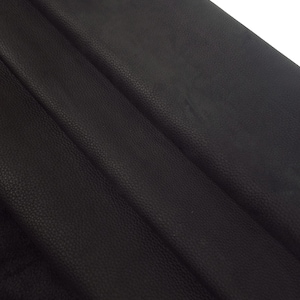 Può includere: Un pezzo di pelle nera caratterizzato da una distinta texture granulata. Il materiale è piegato in diversi strati, creando profonde ombre e riflessi diagonali sulla superficie, che mettono in risalto la finitura opaca e la grana.