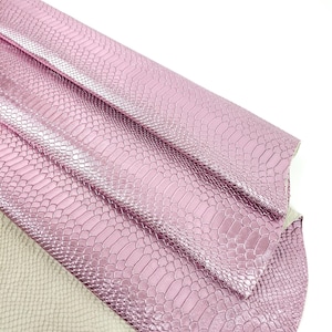 Op de afbeelding: Roze slangenleer geëmbost lederstof. De stof is in drieën gevouwen, waardoor de textuur en kleur van het materiaal zichtbaar zijn.