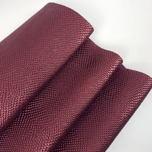 May include: Close-up of a folded sheet of burgundy-colored faux leather. The material has a textured, scaled pattern. The folds reveal the underside of the material, showing a smooth surface. The lighting highlights the sheen of the material.
