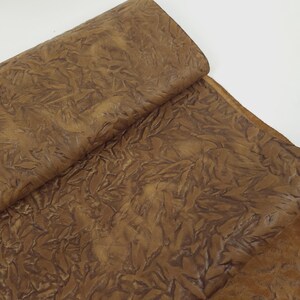 May include: A close-up of a folded piece of brown leather with a textured, leaf-like pattern. The leather has a warm, earthy tone, with variations in color creating depth and dimension. The material appears soft and pliable.