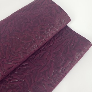May include: Two overlapping pieces of burgundy-colored fabric with a textured, leaf-like pattern. The fabric has a slightly glossy finish, and the folds create shadows that add depth to the design.