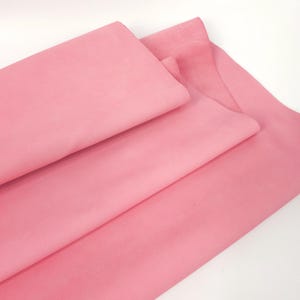 May include: A folded piece of solid light pink fabric with a smooth, matte texture. The material is draped in layers, showing a consistent pastel pink color throughout the surface. The fabric appears soft and pliable, with visible folds and creases.