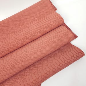 Puede incluir: Primer plano de un trozo de tela de imitación de cuero de color coral doblado con un patrón en relieve de piel de serpiente.