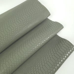 May include: A folded sheet of sage green faux leather material featuring a textured snakeskin pattern. The surface displays a repeating scale design with a matte finish, showing multiple layers of the material stacked in a cascading arrangement.