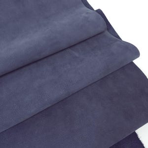 May include: A close-up shot of several layers of navy blue fabric. The fabric has a soft, suede-like texture and is folded over itself. The layers create shadows and depth, highlighting the material's texture and colour.