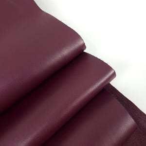 May include: A folded piece of deep burgundy leather with a smooth, matte finish on the top surface and a textured, suede-like appearance on the underside. The material is flexible and shows a consistent dark red colour throughout the folds.