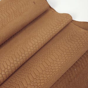 Op de afbeelding: Close-up van bruin leer met een reptielachtig reliëfpatroon. Het leer is gevouwen, waardoor de textuur en kleurvariaties zichtbaar worden. Het materiaal lijkt zacht en soepel, geschikt voor diverse knutselprojecten.