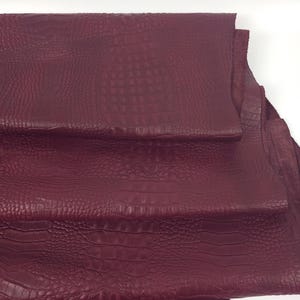 May include: Burgundy-colored leather sheets with a crocodile skin texture. The sheets are stacked, revealing the detailed pattern and color variations. The material appears to be high-quality, suitable for various crafting projects.