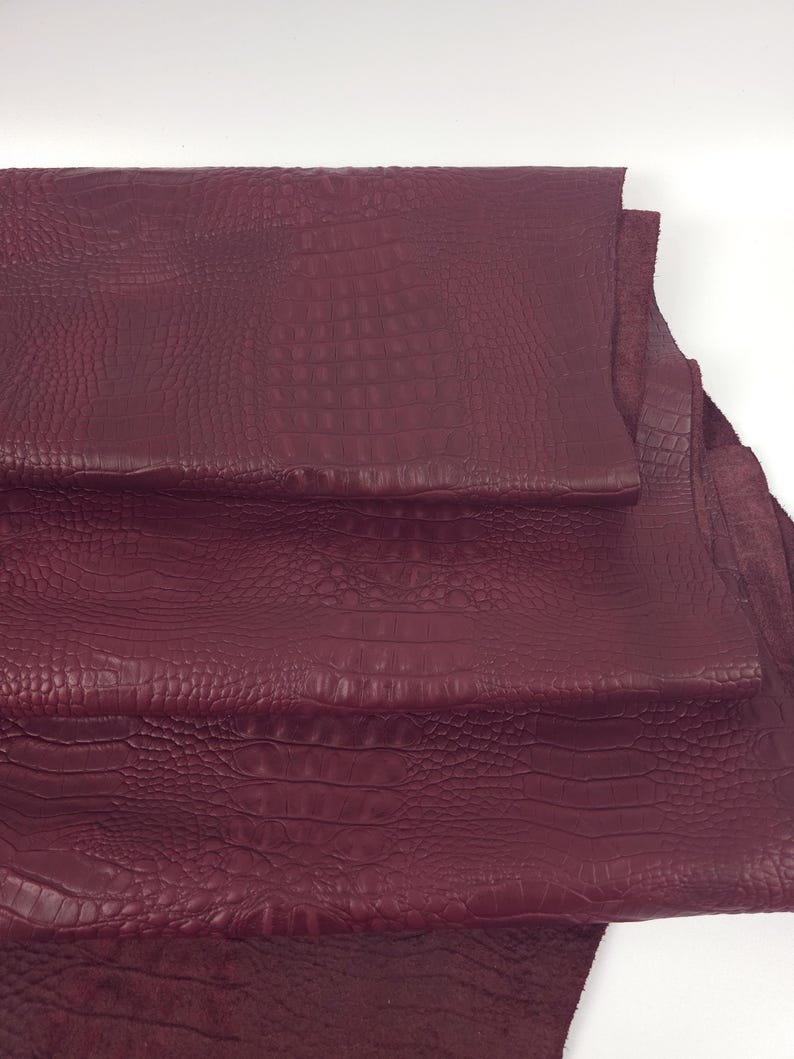 May include: Burgundy-colored leather sheets with a crocodile skin texture. The sheets are stacked, revealing the detailed pattern and color variations. The material appears to be high-quality, suitable for various crafting projects.