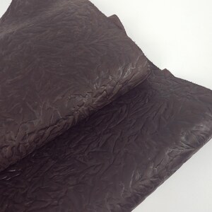 May include: Close-up of a dark brown leather sheet with a textured, wrinkled surface. The leather appears to be folded over itself, revealing the underside. The lighting highlights the variations in the material's texture.