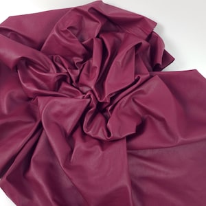 Pode incluir: Um grande pedaço de tecido de couro sintético bordô, artisticamente drapeado e dobrado. O material tem uma superfície lisa e ligeiramente brilhante, com dobras e vincos visíveis que criam sombras. O tecido está sobre um fundo branco.