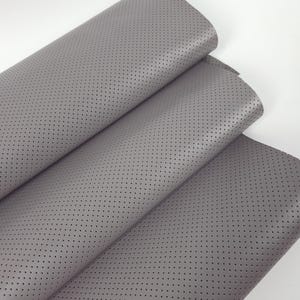 Puede incluir: Primer plano de tela de cuero gris perforada. El material está doblado, revelando múltiples capas. La superficie está salpicada de pequeños agujeros espaciados, creando una apariencia texturizada. La tela tiene un acabado mate y parece suave.