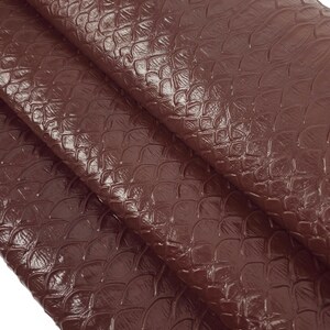 May include: A close-up of a brown leather fabric with a textured, scaly pattern. The fabric is folded in thirds, revealing the texture of the material.