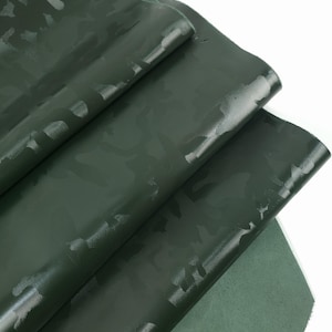 Puede incluir: Tela de cuero verde oscuro con un patrón de camuflaje. La tela está doblada, mostrando la superficie superior brillante y la parte inferior mate. El diseño de camuflaje es de un tono verde más claro, creando un contraste sutil.
