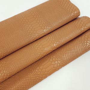 Puede incluir: Primer plano de una pila de cuero sintético marrón con un patrón en relieve de cocodrilo. El cuero está doblado en tres partes, mostrando la textura del material.