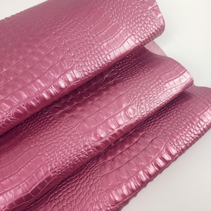 Könnte beinhalten: Ein Bogen aus metallisch rosa Material mit einer erhabenen, geprägten Krokodilleder-Struktur. Das Material ist in drei deutliche Lagen gefaltet und zeigt das sich wiederholende Schuppenmuster sowie die reflektierende, glänzende Oberfläche.