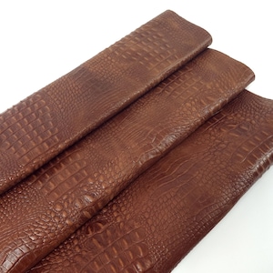 May include: Close-up of three pieces of brown faux crocodile leather. The material has a textured, embossed pattern that mimics the look of crocodile skin. The leather is a warm, medium brown color.
