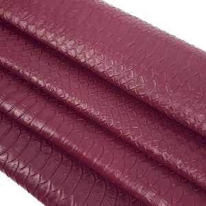 Könnte beinhalten: Nahaufnahme eines burgunderfarbenen Kunstleders mit Schlangenhautmuster. Der Stoff ist gefaltet und hat eine strukturierte, schuppige Textur.